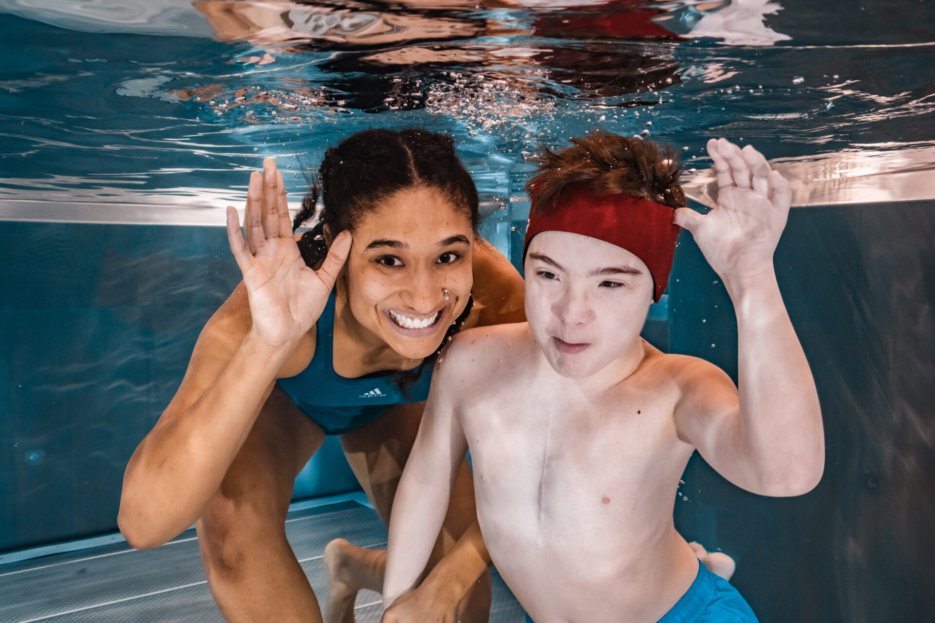 Zwei Personen unter Wasser in einem Schwimmbad, die lächeln und in die Kamera winken. Die Person auf der linken Seite hat dunkles Haar in Zöpfen und trägt einen blauen Badeanzug, während die Person auf der rechten Seite kurze Haare hat und ein rotes Stirnband und eine blaue Badehose trägt.